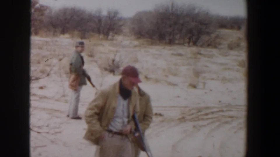 A red-capped hunter with a rifle walks past the camera while another adult hunter stands in the brushy Lubbock field behind him.