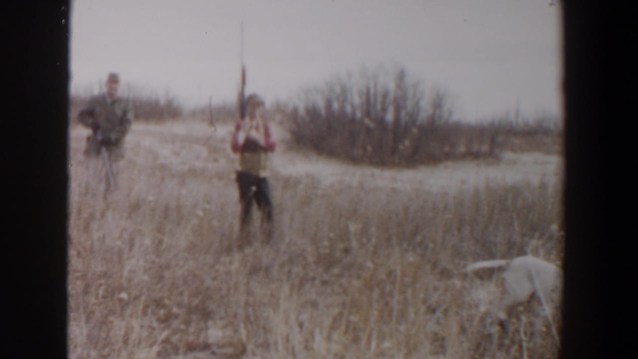 Two men with rifles stand in a dry field while a white dog moves through tall grass.