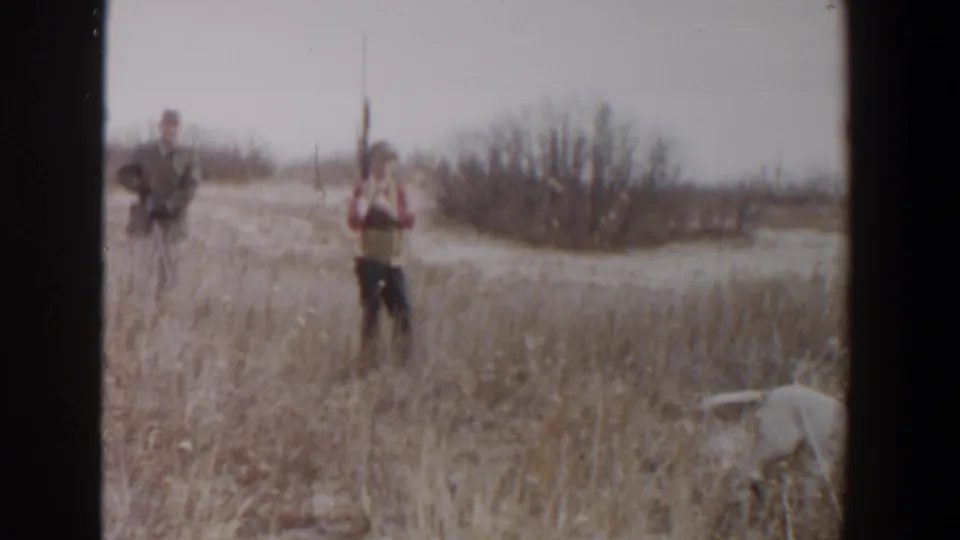 In 1956 Lubbock, Texas, two adult men stand with rifles in a dry field as a white dog moves through the foreground grass.
