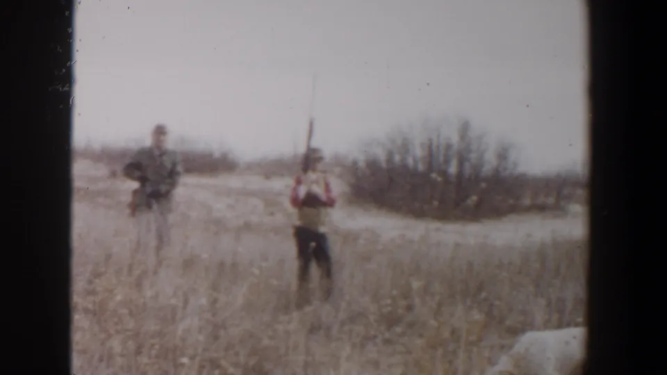 Two adult men with rifles stand in a dry Lubbock field, with brush behind them and a white dog partly visible at the lower right.
