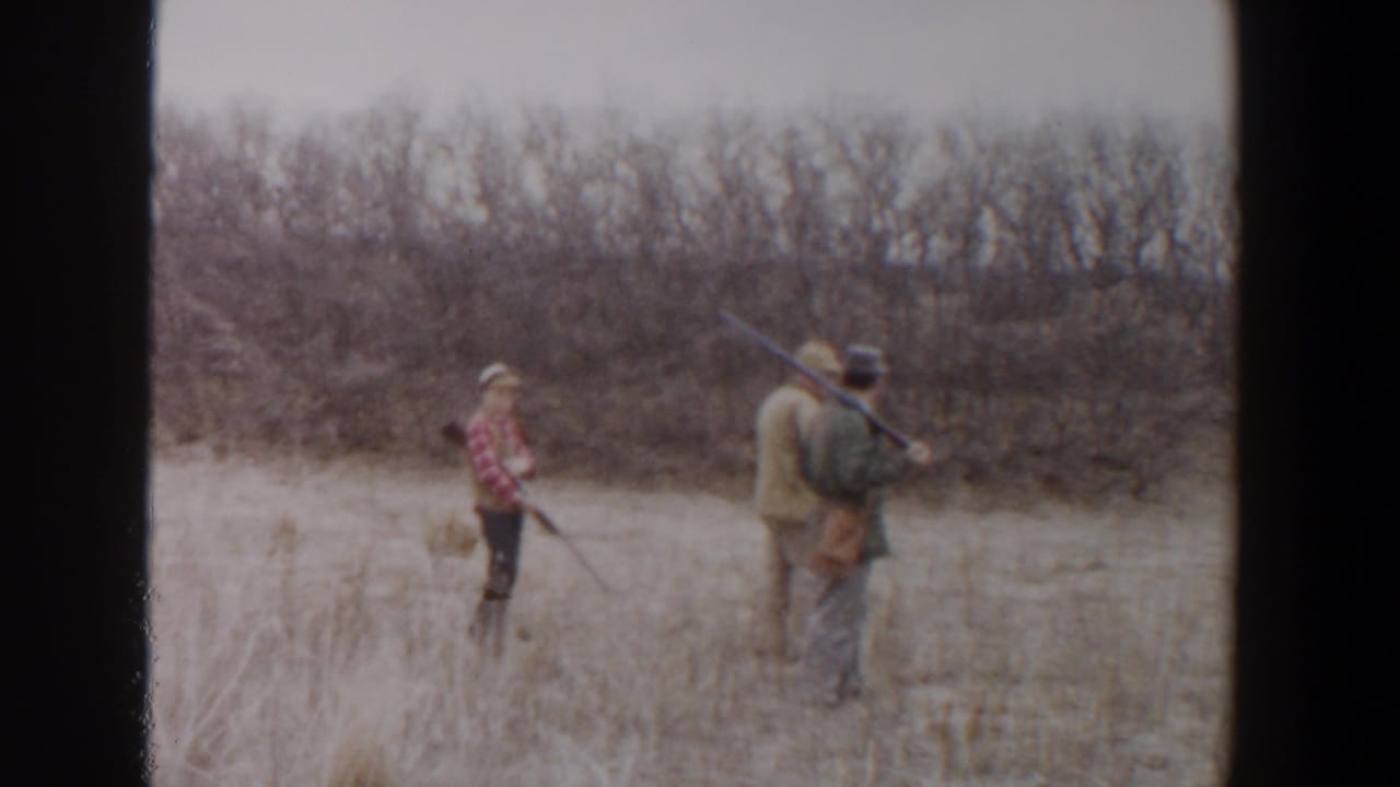 1956 Lubbock, Texas home-movie frame showing three people with rifles crossing dry grass near a brushy treeline.