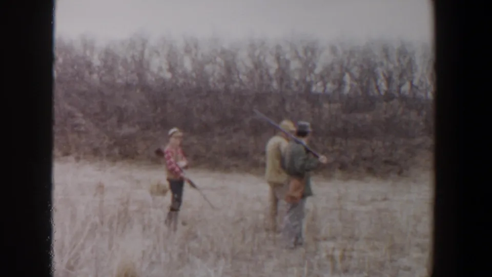1956 Lubbock, Texas home-movie frame showing three people with rifles crossing dry grass near a brushy treeline.