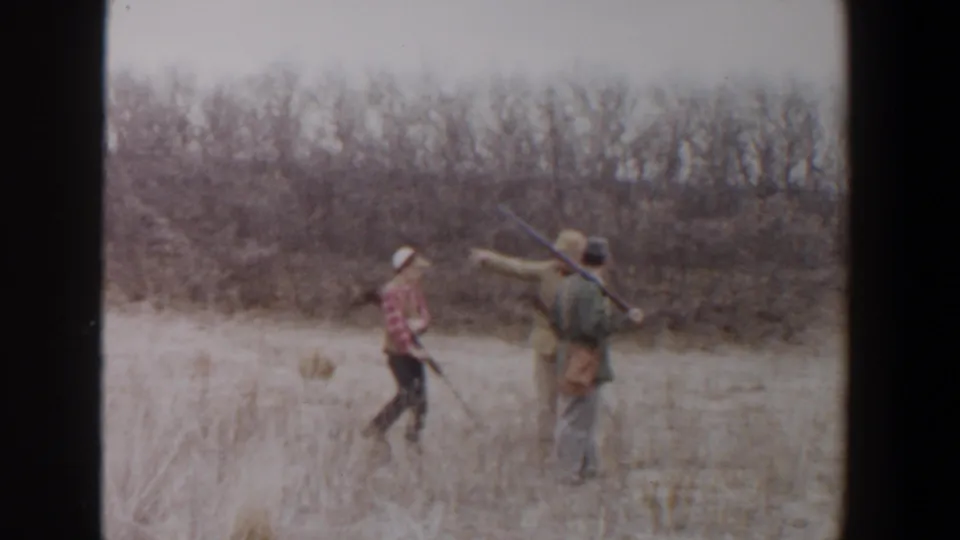 1956 Lubbock, Texas frame of three people walking with rifles through tall grass, with one adult gesturing toward brush.