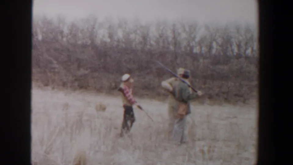 Home-movie frame from 1956 Lubbock, Texas showing three people with rifles moving away through dry brush land.