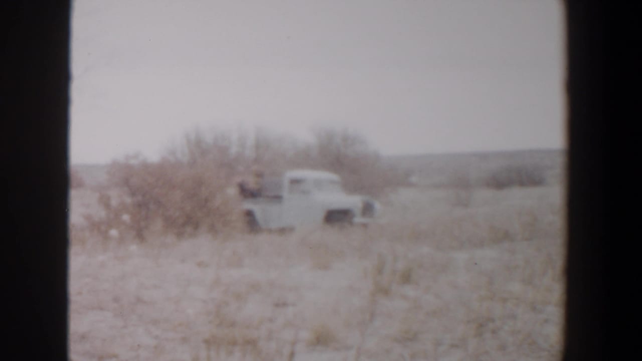 1956 Lubbock, Texas 8mm frame showing a white pickup moving through dry countryside with people riding in the open bed.