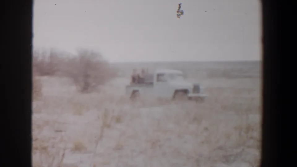 Faded 8mm frame of a white pickup crossing dry open land near Lubbock, Texas, with two visible riders in the truck bed.
