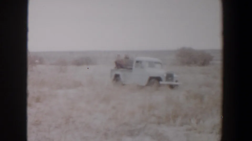 1956 Lubbock-area home-movie frame showing a white pickup crossing dry grassland with two riders in the open bed.