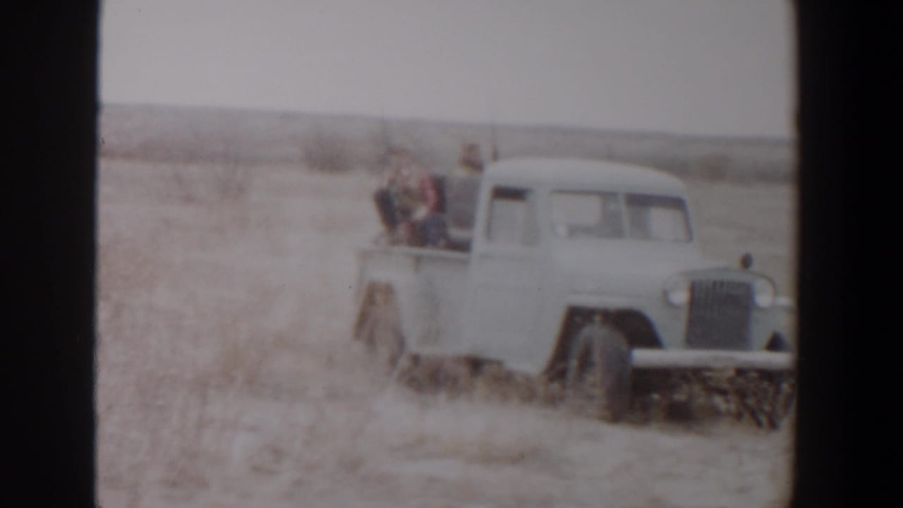 White pickup crossing a dry field with men riding in the truck bed.