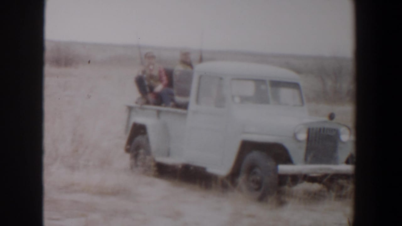 White pickup in an open field with two men seated in the rear.