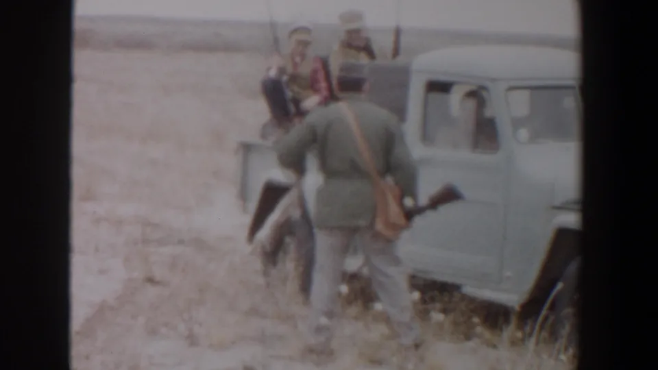 A man with a rifle and game animal appears near the white pickup, with two men seated in the bed in the Texas field.