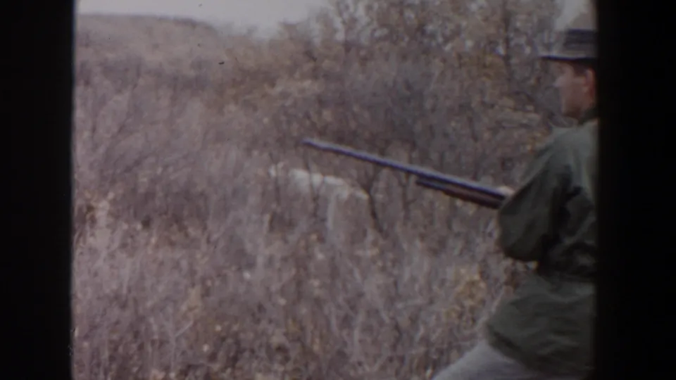 In the supplied 1956 Lubbock footage, a man in a green jacket and hat moves through dry brush with a rifle.