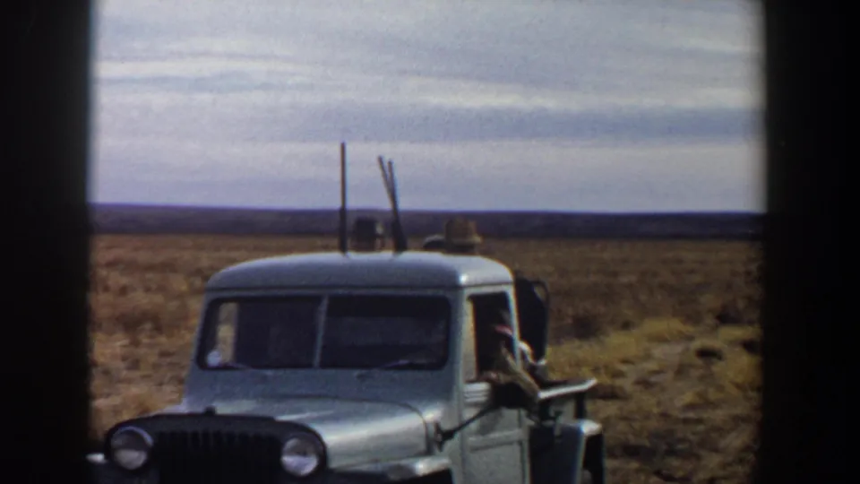 A pale pickup moves through open country near Lubbock, Texas, with adult men and upright rifles visible in the rear.