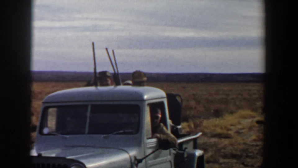 The front of a light pickup fills the frame while adult men sit behind the cab with rifles held upright in the 1956 Texas scene.
