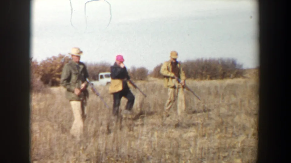 Three men in hunting clothes walk through a dry field near Lubbock, Texas, each carrying a rifle, with a light vehicle in the background.
