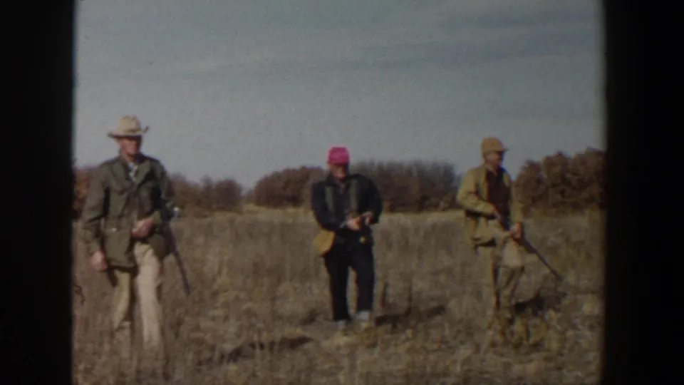 In this 1950s Texas home-movie frame, three hunters are spaced across a dry field with guns lowered.