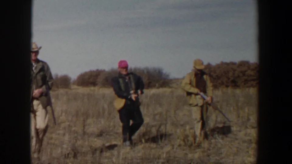 Three adult hunters near Lubbock walk through dry grass, with hats, long guns, and brush visible behind them.