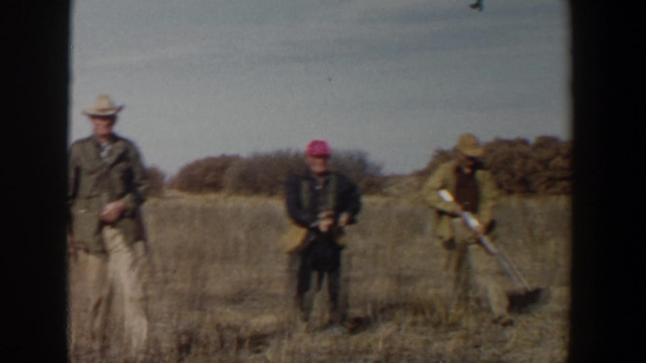 Three adult men with rifles stand in a dry grassy field near low brush.