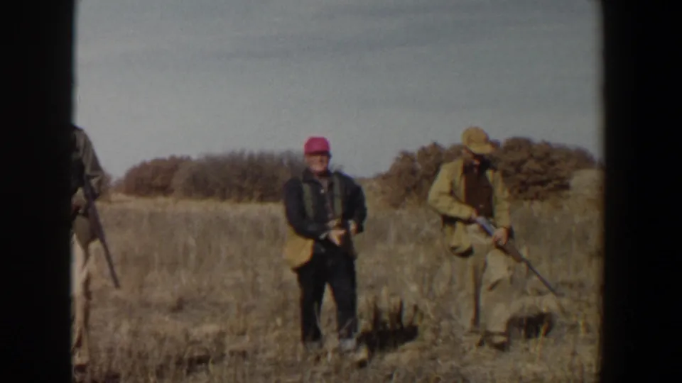 Two adult men in hunting attire carry rifles through a dry Texas field, with a partial third figure near the frame edge.