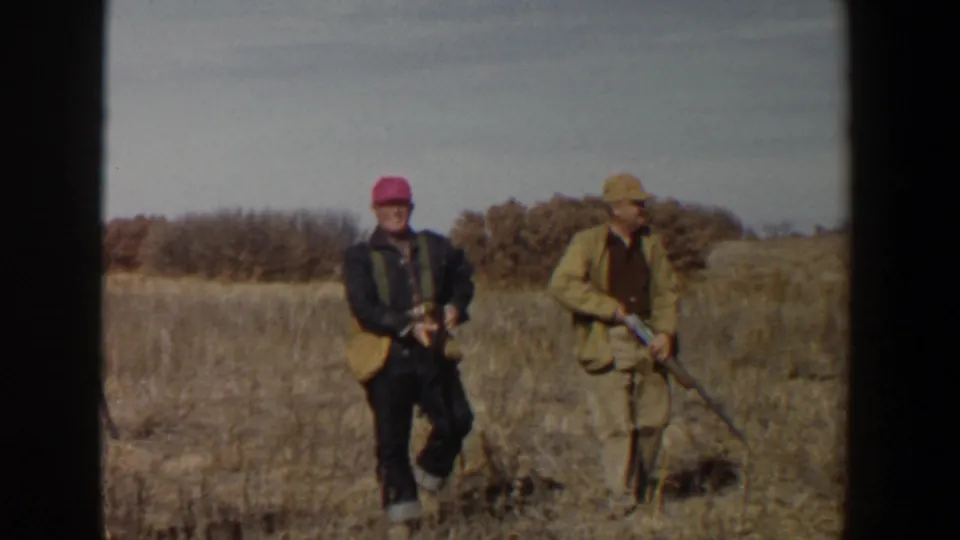 Two adult hunters in 1956 Lubbock, Texas footage walk across dry grass, one wearing a bright pink cap and the other a tan hat.