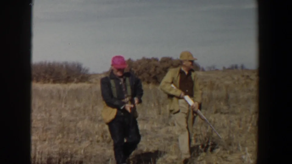 Two adult men in vintage hunting clothes walk with rifles through a dry field in the Lubbock, Texas home-movie scene.