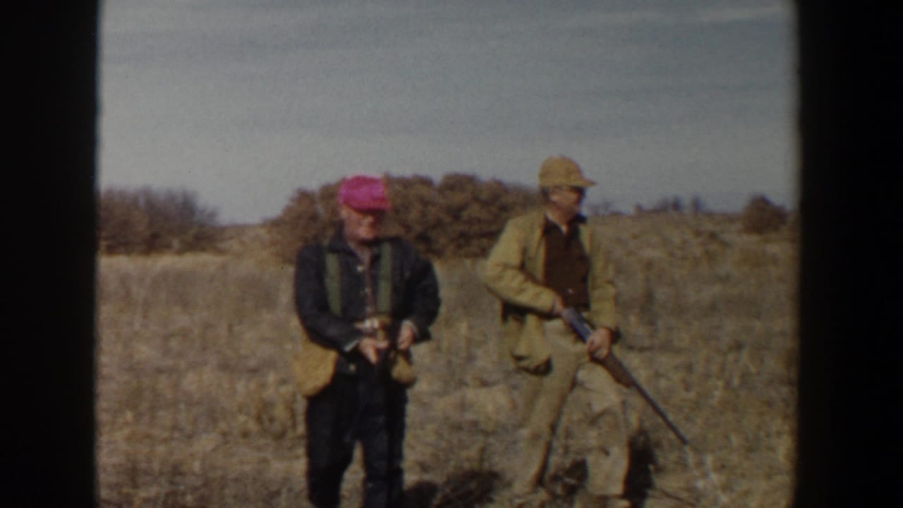 Two adult hunters stand in dry grass holding rifles, one wearing a bright cap and the other a light hat.