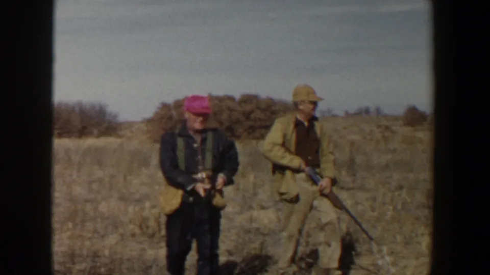 Two adult hunters in 1950s Lubbock, Texas countryside stand in dry grass with rifles, filmed in soft faded 8mm color.