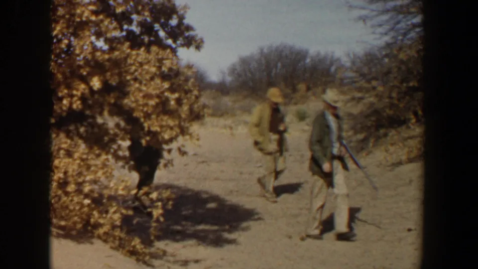 Two hunters in 1950s Lubbock, Texas countryside walk along a dusty path with rifles, framed by dry brush and trees.