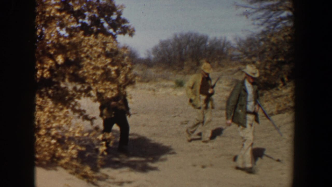 Three men with rifles walk across a dry field bordered by brush and bare trees.