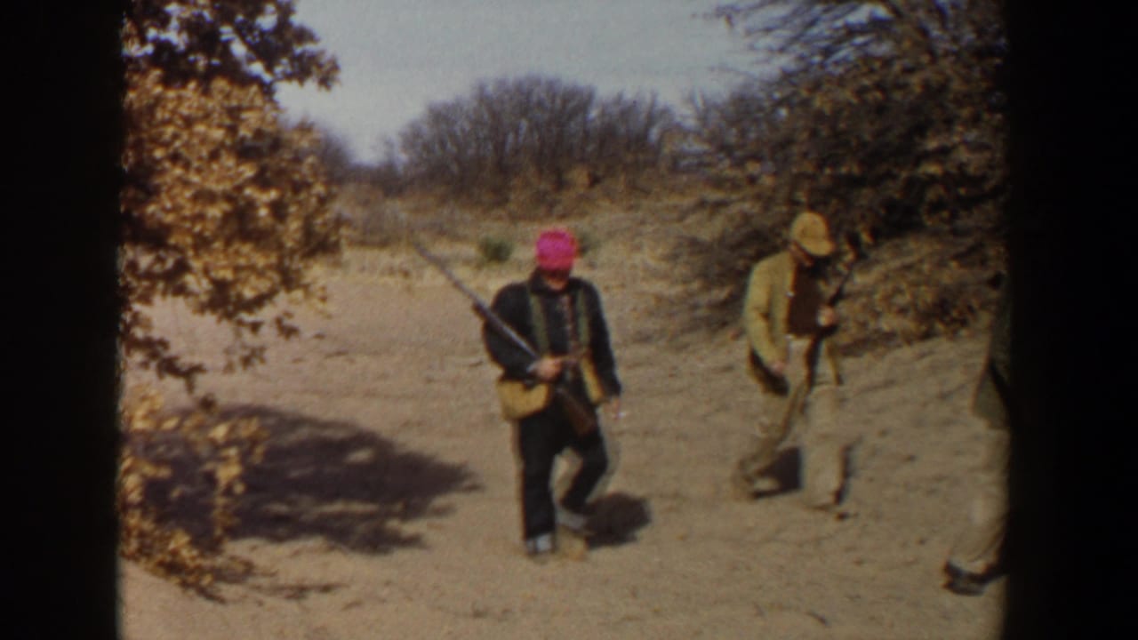 The home-movie frame shows rifle-carrying hunters moving across sandy West Texas terrain with brush and bare trees behind them.