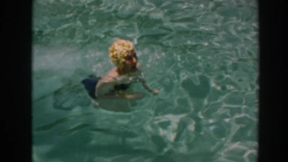 The woman surfaces in the 1961 North Hollywood backyard pool, her blonde hair or cap visible as she swims toward the camera.