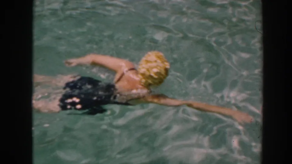 In 1961 North Hollywood home-movie footage, a woman in a yellow swim cap and black swimsuit swims through a backyard pool.
