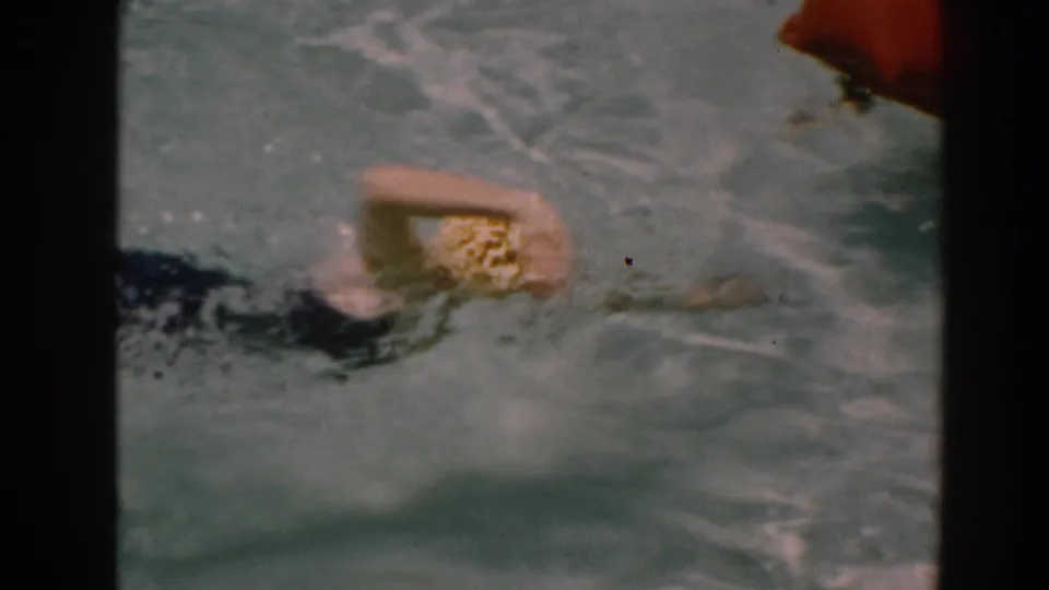 A woman in a yellow swim cap continues swimming in a backyard pool, seen in soft 8mm color footage from North Hollywood in 1961.