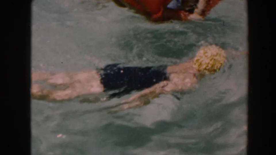 The swimmer in a yellow cap and black swimsuit moves near the edge of a backyard pool in 1961 North Hollywood footage.