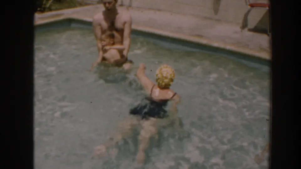 A woman in a yellow swim cap swims toward a seated man at a backyard pool edge in North Hollywood, California, in 1961.