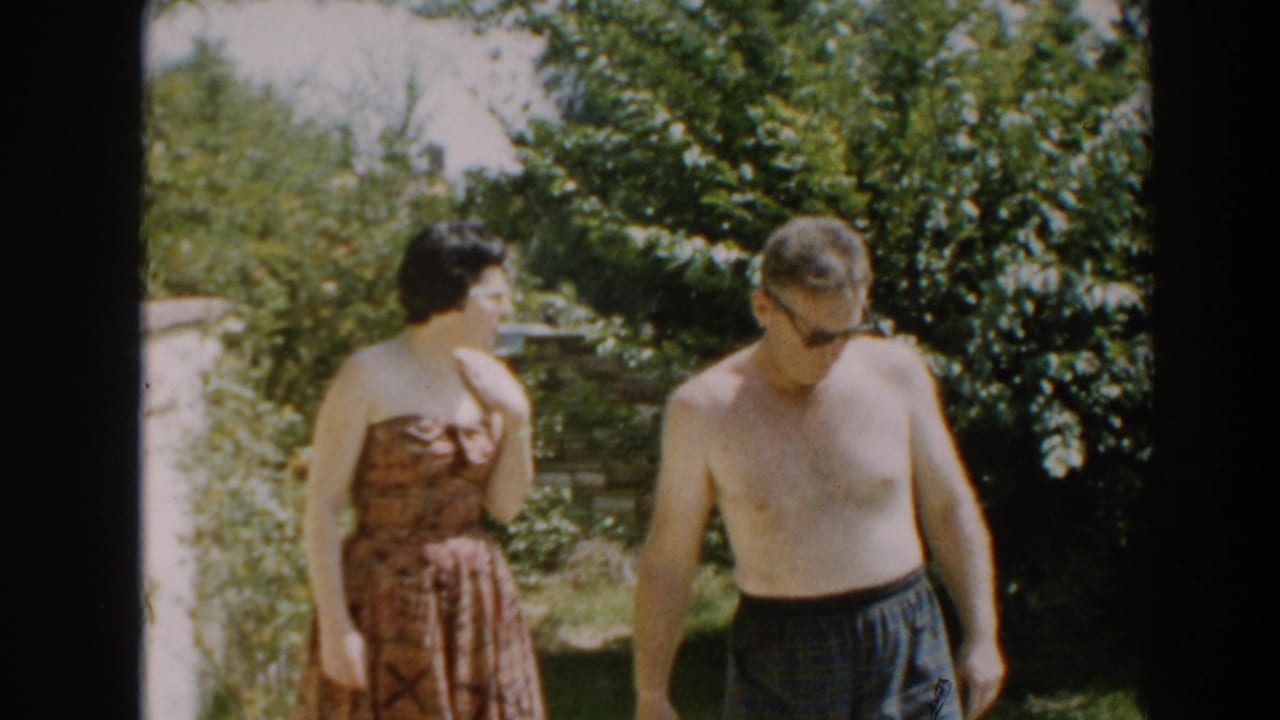 A shirtless man and a woman in a strapless dress walk through a sunny backyard.