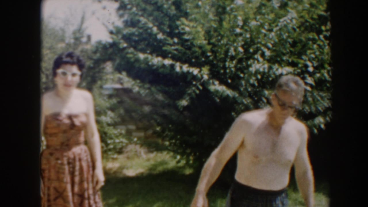The woman faces the camera while the shirtless man walks at the right edge of the yard.