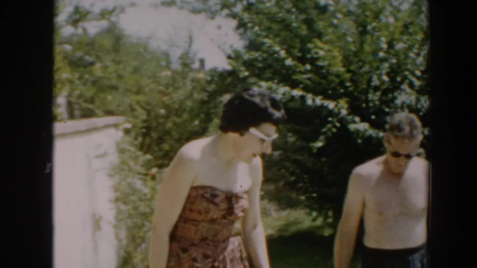 In a 1961 North Hollywood backyard, a woman in white sunglasses stands near a shirtless man wearing dark sunglasses and plaid swim trunks.