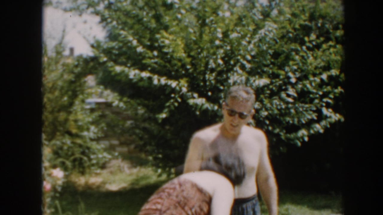 Woman bends forward in the foreground while a shirtless man stands in the backyard.