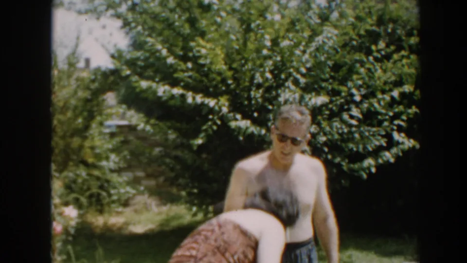 The woman bends down at the edge of the frame as the shirtless man in sunglasses stands beside garden greenery in North Hollywood.