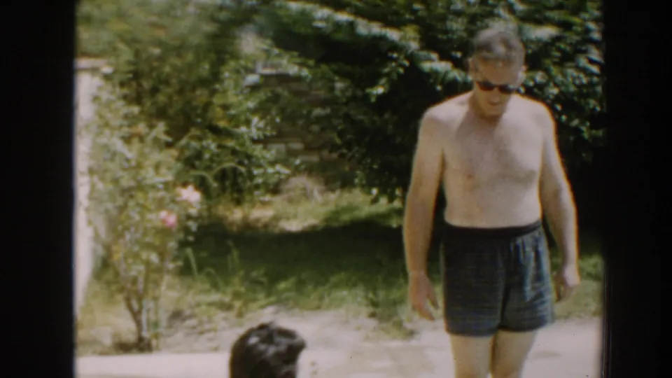 A man in dark sunglasses and plaid swim trunks stands in the bright 1961 backyard while the woman is partly visible near the lower edge.