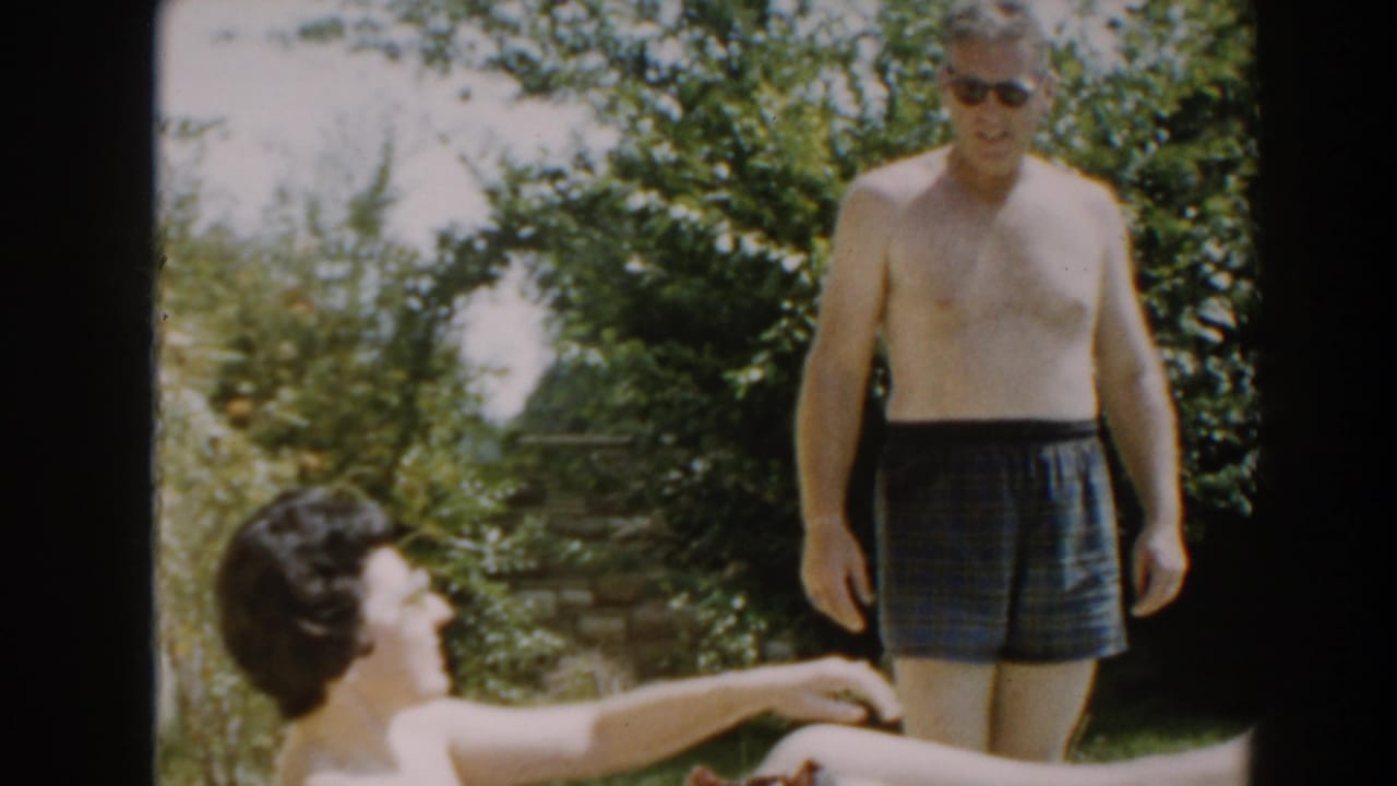 1961 North Hollywood home-movie frame of a woman reclining in a backyard as a shirtless man in dark swim trunks stands near shrubs.
