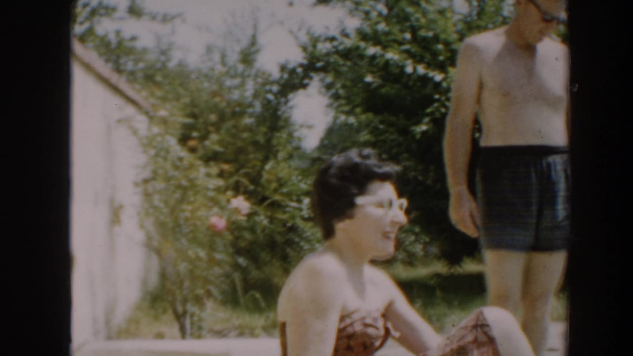 Woman in white sunglasses sits smiling while a shirtless man stands at the right edge.
