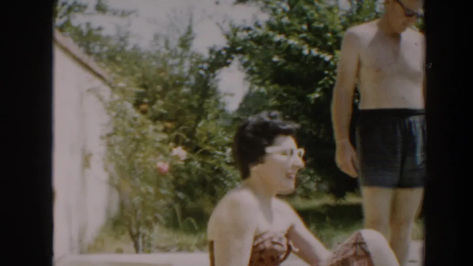 A woman in white-framed sunglasses and a patterned swimsuit smiles in a 1961 North Hollywood backyard as a man stands nearby.