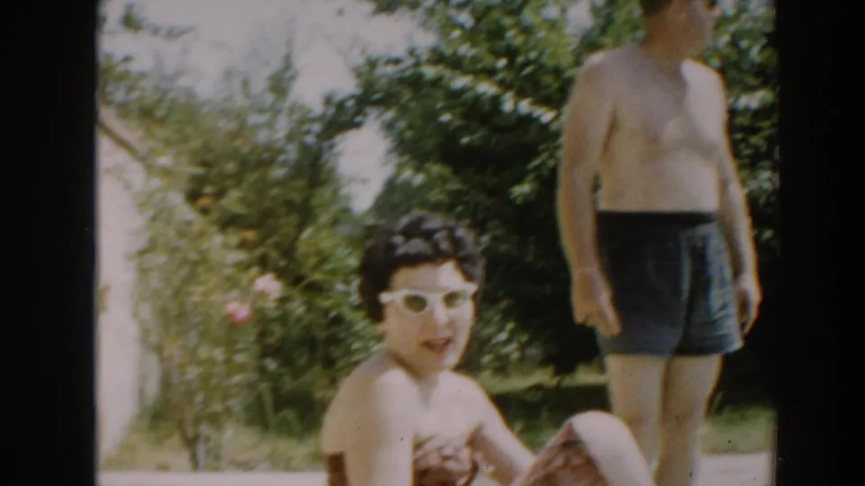 Backyard home-movie frame showing a woman in white sunglasses looking toward camera with a shirtless man behind her in swim trunks.