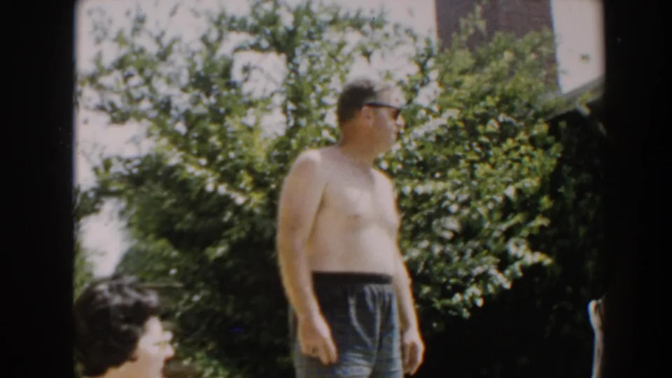 1961 North Hollywood backyard frame with a shirtless man in dark swim trunks standing in profile beside a seated woman and garden shrubs.