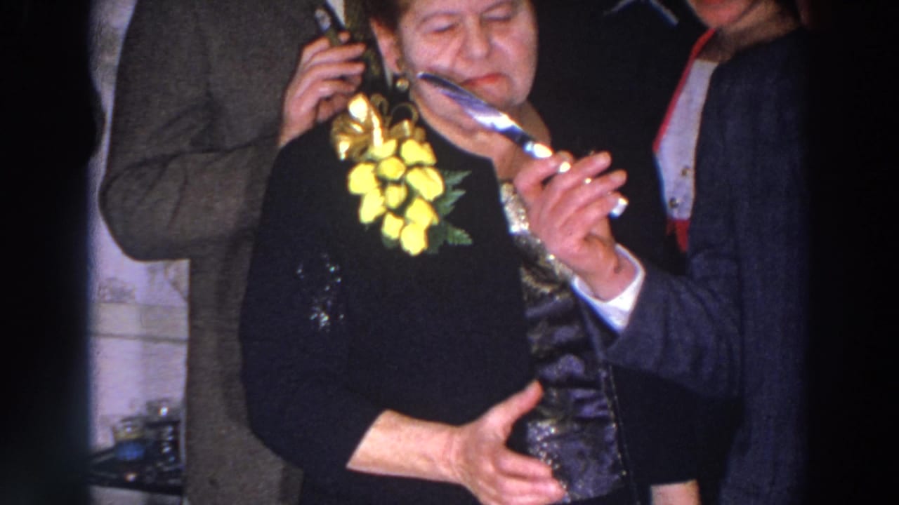 Older woman with yellow corsage holds a knife while a suited man assists.