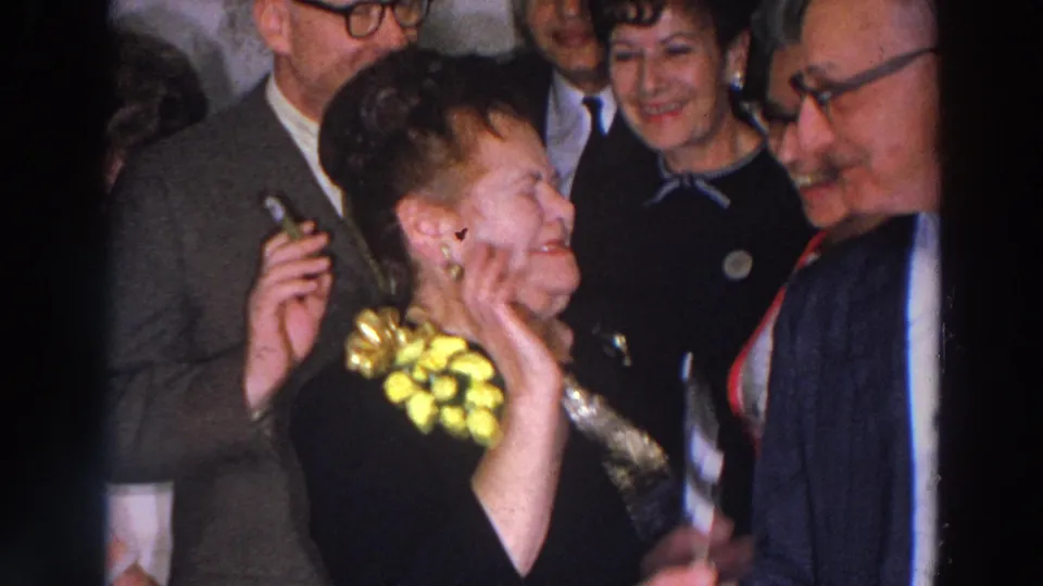 1966 Madison home-movie frame showing the corsage-wearing woman smiling as guests in suits and dresses gather close around her.