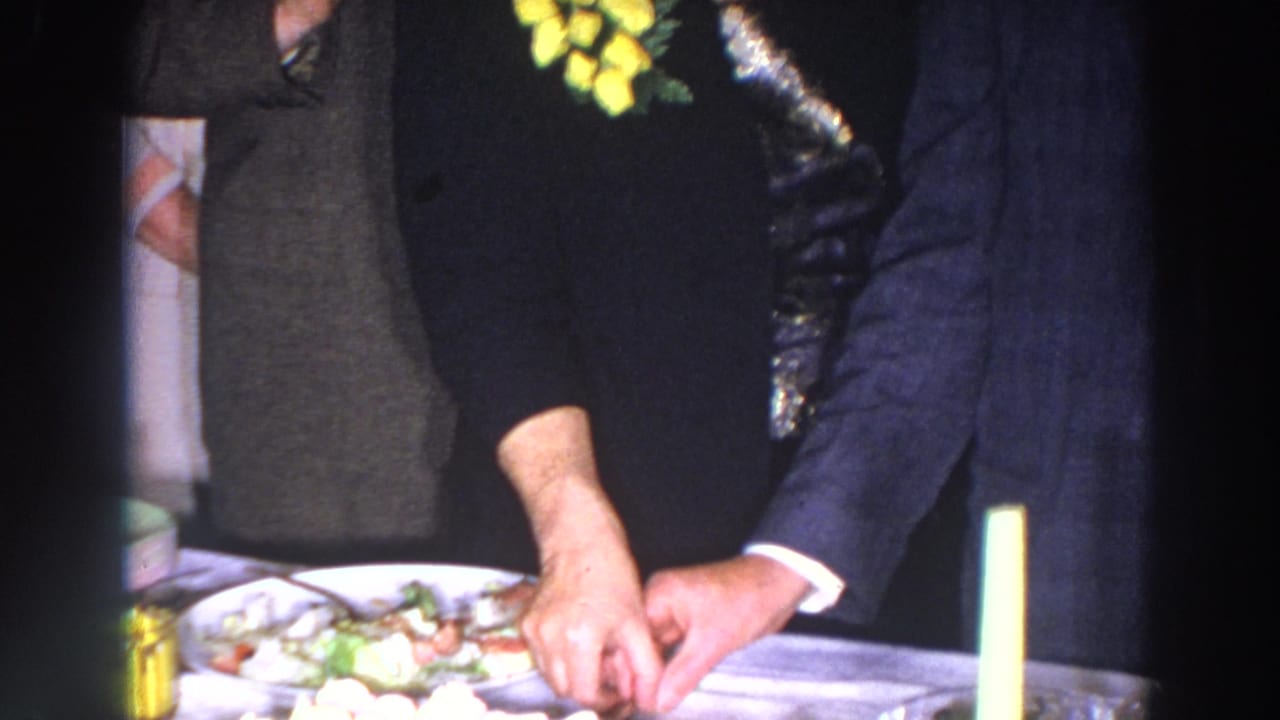 Hands cut cake on a table as formally dressed adults stand nearby.