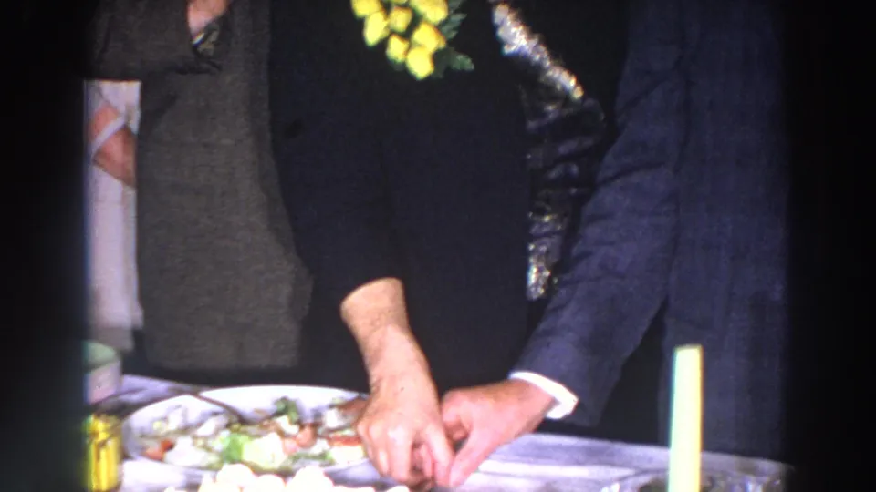 Madison 1966 home-movie frame showing a cake table with adult guests in suits and formal clothing gathered around it.