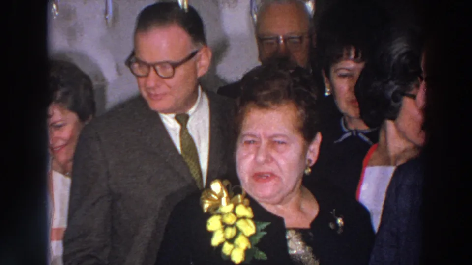 Madison 1966 party frame with adult guests in suits, glasses, formal dresses, and a prominent yellow corsage in a crowded interior.